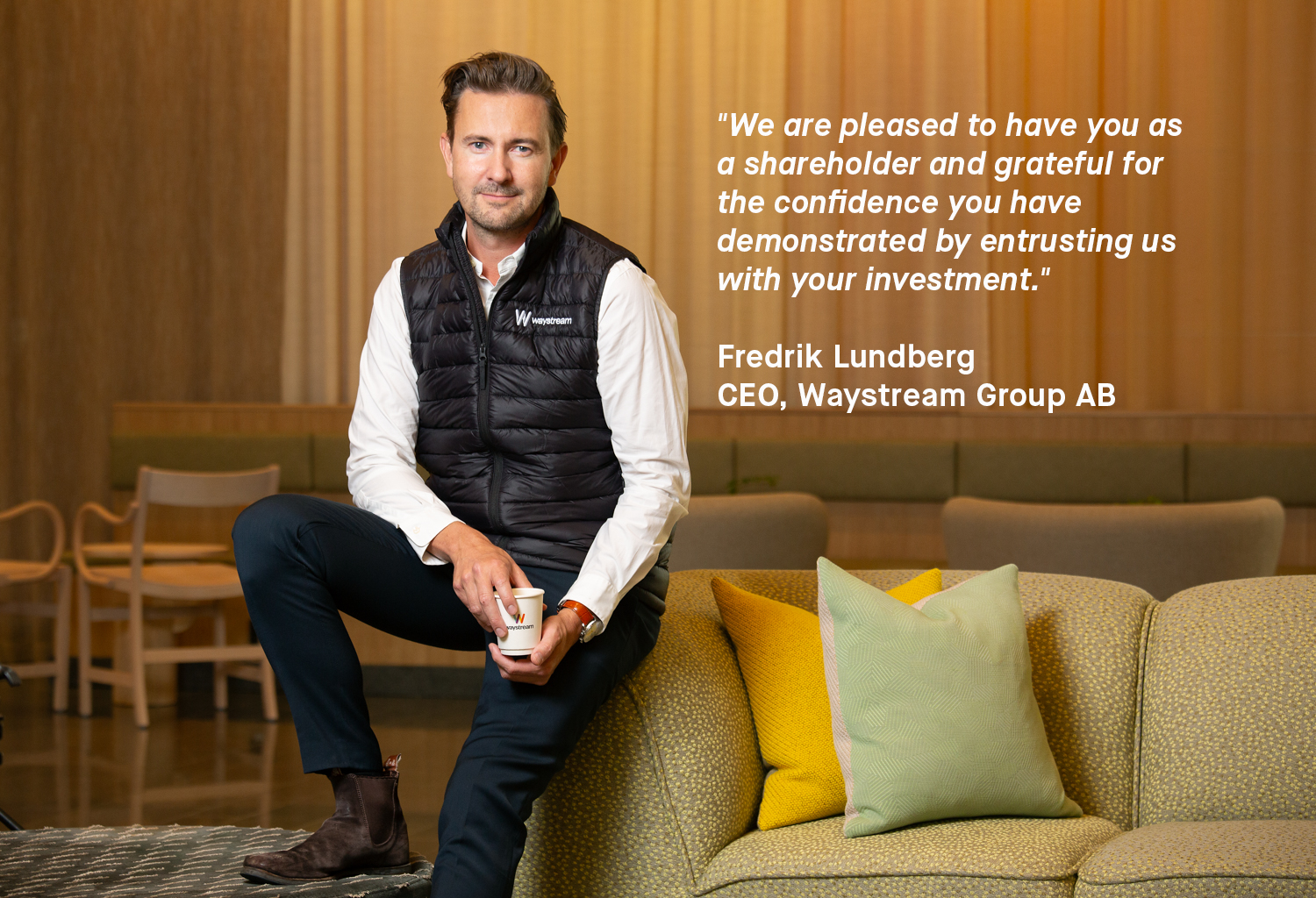 Person sitting on a sofa holding a coffee cup, wearing a white shirt and black Waystream vest with colorful cushions nearby.