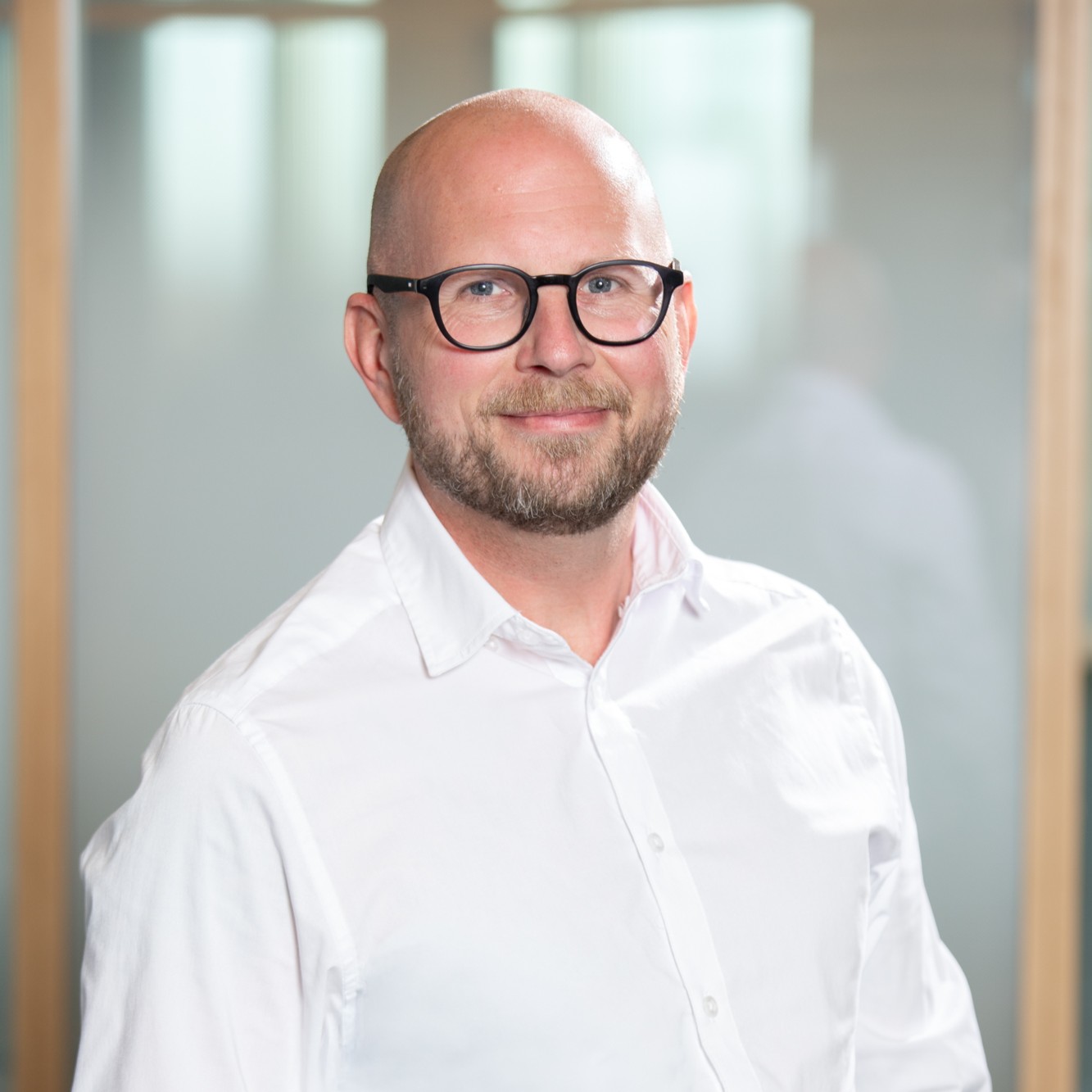 En person med runda svarta glasögon och en vit skjorta mot en svagt suddig kontorsbakgrund.
